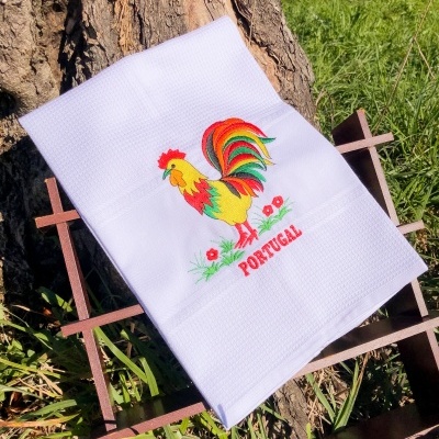 Toalha branca com galo colorido bordado e a palavra PORTUGAL junto a tronco de árvore
