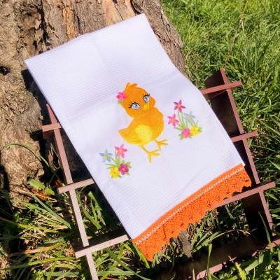Toalha branca com bordado de pintainho amarelo e flores, com borda de croché laranja, sobre suporte de madeira ao ar livre