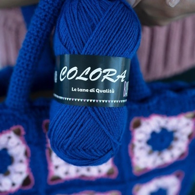 Novelo de lã azul com etiqueta preta da marca Colora e peça de crochet azul e rosa ao fundo
