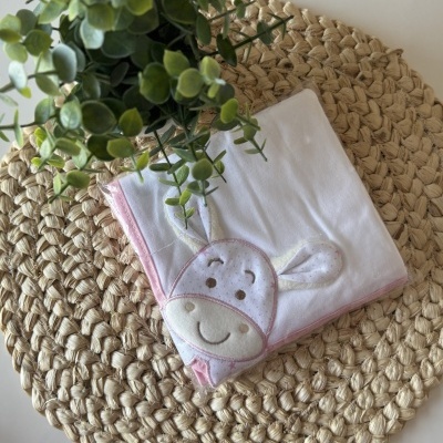 Manta branca com bordas rosa e desenho de crocodilo, sobre suporte redondo de fibra trançada e planta verde