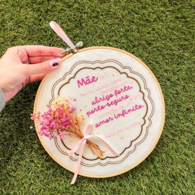 Aro de bordado com mensagem bordada e flores secas amarradas por fita rosa, mostrado sobre relva.