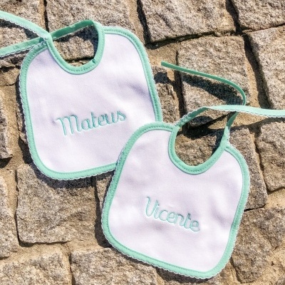Dois babetes brancos com bordas verdes e nomes bordados colocados num chão de pedras