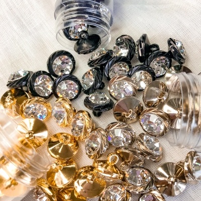 Botões decorativos dourados, prateados e pretos com cristais transparentes no centro, espalhados sobre tecido branco e frascos plásticos