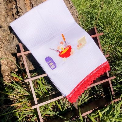 Toalha branca texturizada com bordado colorido e borda vermelha numa estrutura de madeira junto a uma árvore.