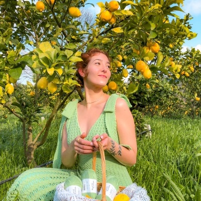 Mulher vestindo vestido verde de crochê segurando cesta com garrafas e limões perto de árvore de limão