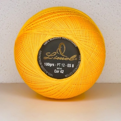 Novelo de linha amarela em algodão mercerizado com etiqueta preta e dourada, sobre superfície clara.