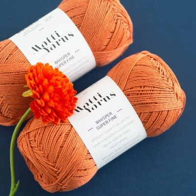 Dois novelos de lã laranja Whisper Super Fine com rótulos brancos e uma flor laranja ao lado sobre fundo azul.
