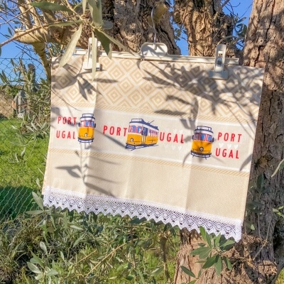 Pano de cozinha bege com estampas de elétricos amarelos e texto 'PORTUGAL', pendurado numa árvore