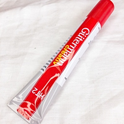 Tubo de cola metálico com tampa vermelha e texto 'Gütermann creativ HT2' sobre tecido branco