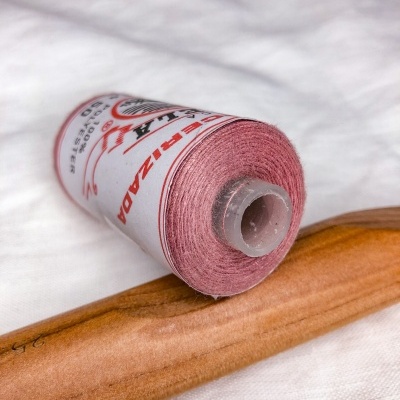 Rolo de linha cor-de-rosa com rótulo e agulha de madeira sobre tecido branco.