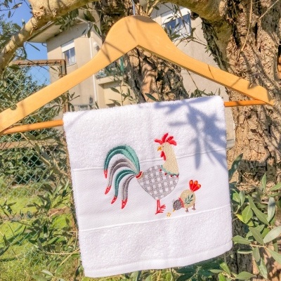 Toalha branca com bordado de galo e pintainho pendurada num cabide de madeira numa árvore