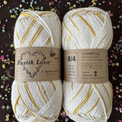 Dois novelos de lã branca e amarela Rustik Love com etiquetas de papel kraft sobre fundo escuro com flores secas coloridas.