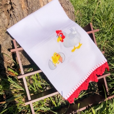 Toalha branca com bordado de galinha e pintainho, com borda em croché vermelho, sobre grelha de madeira no jardim