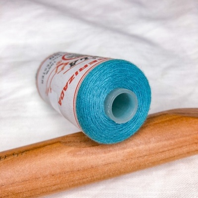 Rolo de linha azul turquesa com rótulo branco e vermelho, colocado sobre superfície de madeira castanha clara.