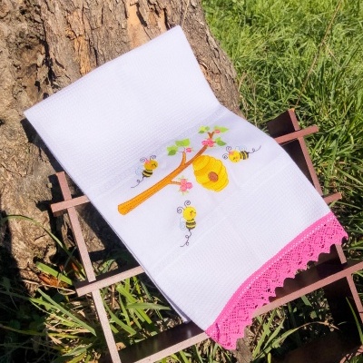 Toalha branca bordada com colmeia e abelhas, com barra rosa, sobre estrutura de madeira na relva
