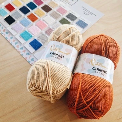 Novelos de lã bege e laranja com etiquetas 'CARINHO Anti-Pilling' sobre madeira clara com tabela de cores ao fundo