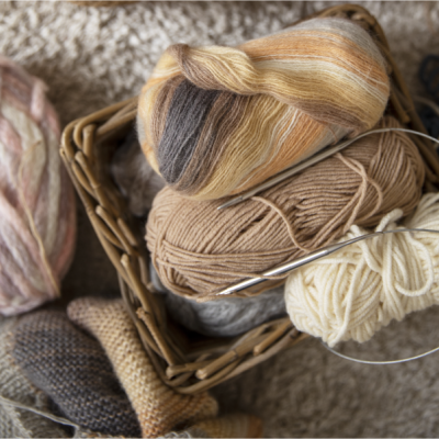 Novelos de lã de tons castanhos e bege com agulhas de tricô dentro de um cesto de vime sobre carpete