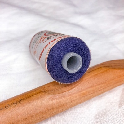 Rolo de linha roxa com rótulo em tecido branco junto de alfinete de madeira.