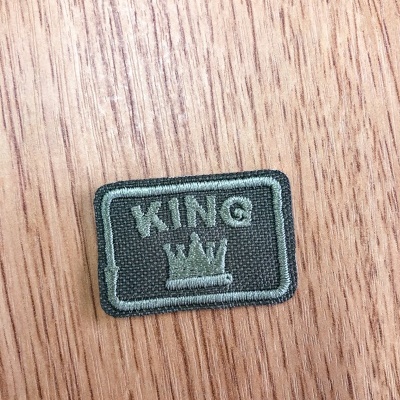 Emblema bordado cinza escuro com texto KING e coroa sobre madeira clara