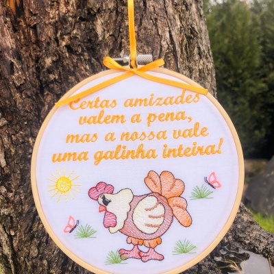 Bastidor bordado com texto e desenho de galinha pendurado numa árvore