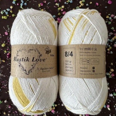 Dois novelos de lã Rustik Love branco e amarelo com etiquetas castanhas, sobre fundo escuro com flores secas coloridas.