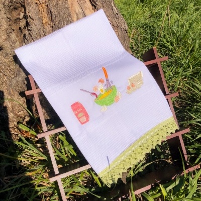 Pano de cozinha branco com bordado de tigela e renda verde clara sobre suporte de madeira ao ar livre