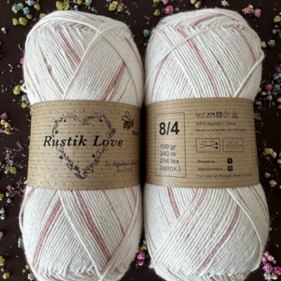 Duas meadas de lã branca com riscas rosas e rótulo castanho Rustik Love sobre fundo escuro com pequenos corpos coloridos.