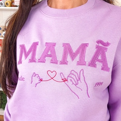 Sweatshirt lilás bordada com palavra MAMÃ e desenho de mãos unidas por fio vermelho em forma de coração.