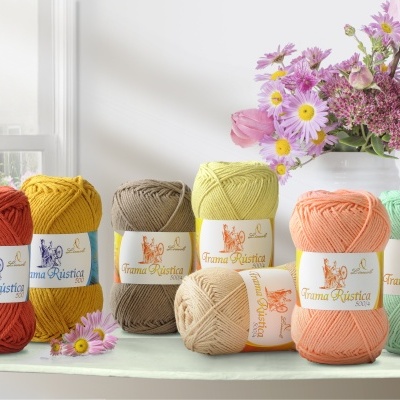 Novelos de lã coloridos da marca Trama Paulista sobre uma mesa com flores ao fundo.