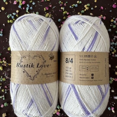 Duas meadas de lã Rustik Love branco com detalhes lilás e rótulos castanhos, sobre fundo com flores coloridas.