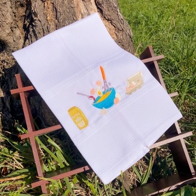 Toalha branca de cozinha com bordado colorido sobre estrutura de metal e madeira junto a tronco de árvore e relva