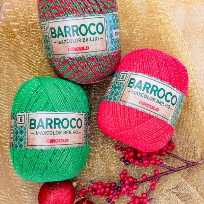 Três novelos de fio colorido brilhoso com etiquetas BARROCO sobre tecido dourado com decoração natalícia.