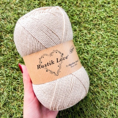Novelo de fio bege claro com rótulo Rustik Love e fundo de relva verde