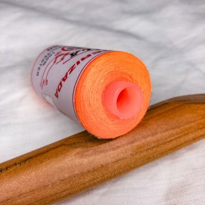 Rolo de linha laranja com bobina de plástico sobre tecido branco e objeto de madeira