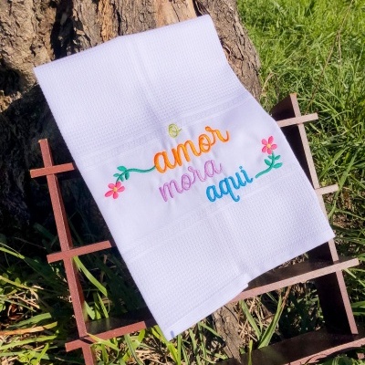 Toalha branca com bordado colorido 'amor mora aqui' em estrutura de ferro junto a tronco e relva