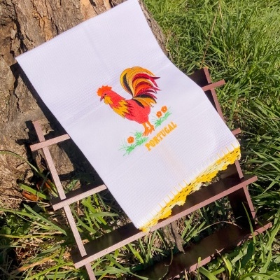 Toalha branca bordada com galo colorido e texto AMOR PORTUGAL em fundo natural