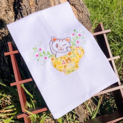 Toalha branca bordada com gato numa chávena amarela, em exposição ao ar livre num suporte de madeira.