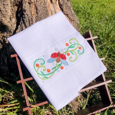 Toalha branca com bordado de joaninha, folhas e flores sobre estrutura de madeira, ao ar livre