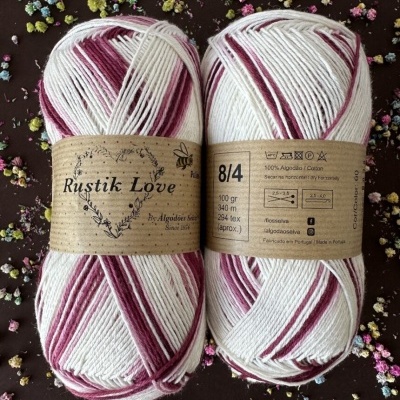 Dois novelos de fio branco e bordô com rótulo Rustik Love de algodão, sobre fundo escuro com flores pequenas coloridas.