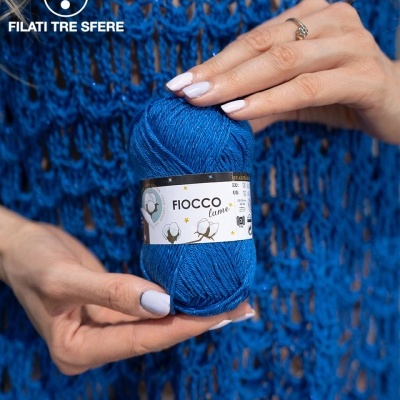 Novelo de fio azul Fiocco lame nas mãos de uma pessoa com suéter azul e logotipo TRE SFERE no fundo