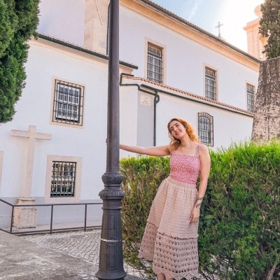 Mulher com vestido floral vermelho e saia bege em exterior junto a poste e edifício branco.