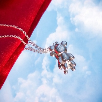 Colar prateado com pendente de urso sobre tecido vermelho e fundo de céu com nuvens