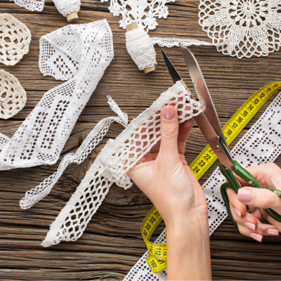 Mãos cortando renda branca com tesoura sobre mesa de madeira com rendas brancas e fita métrica