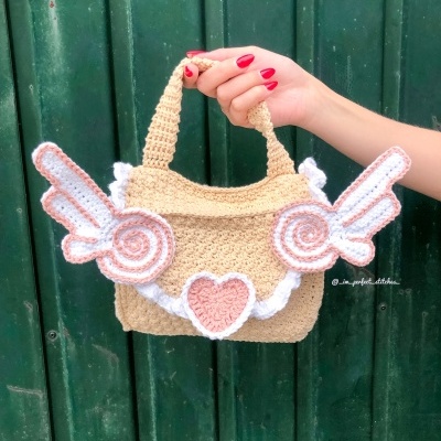 Mala de mão em crochet bege com asas e coração em rosa e branco contra porta verde.