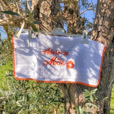 Toalha branca bordada a laranja com texto 'Adoro-te Mãe' pendurada num tronco de árvore