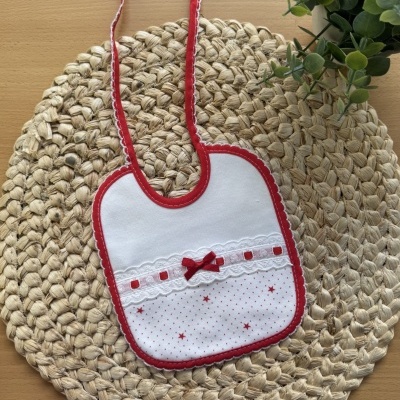 Babete infantil branco com bordas vermelhas e laço, sobre base de palha com planta verde