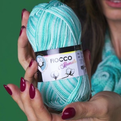 Novelo de fio de algodão azul e branco com etiqueta Fiocco Sfumato, segurado por mulher com unhas vermelhas.