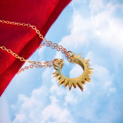 Colar dourado com pendente em forma de sol sobre fundo vermelho e céu azul