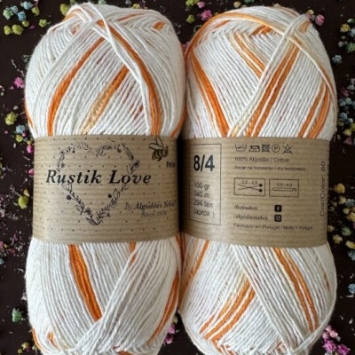 Dois novelos de fio de algodão branco e laranja com etiquetas Rustik Love sobre fundo escuro com flores coloridas.