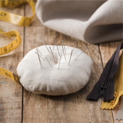 Alfineteiro branco redondo com alfinetes, fita métrica amarela, tecido cinzento e fechos éclair sobre mesa de madeira.